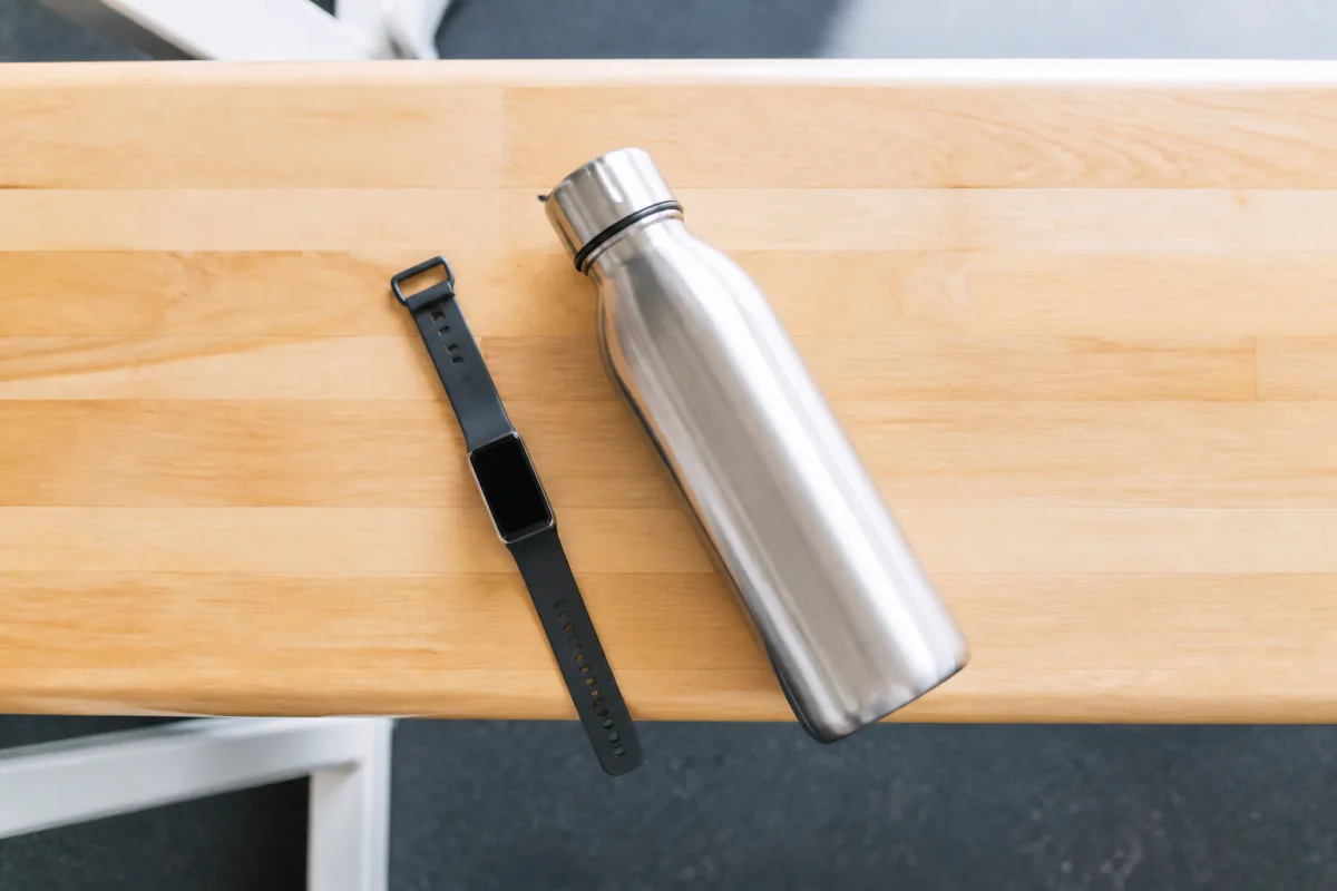 A fitness tracker watch and a water bottle on a bright gym bench