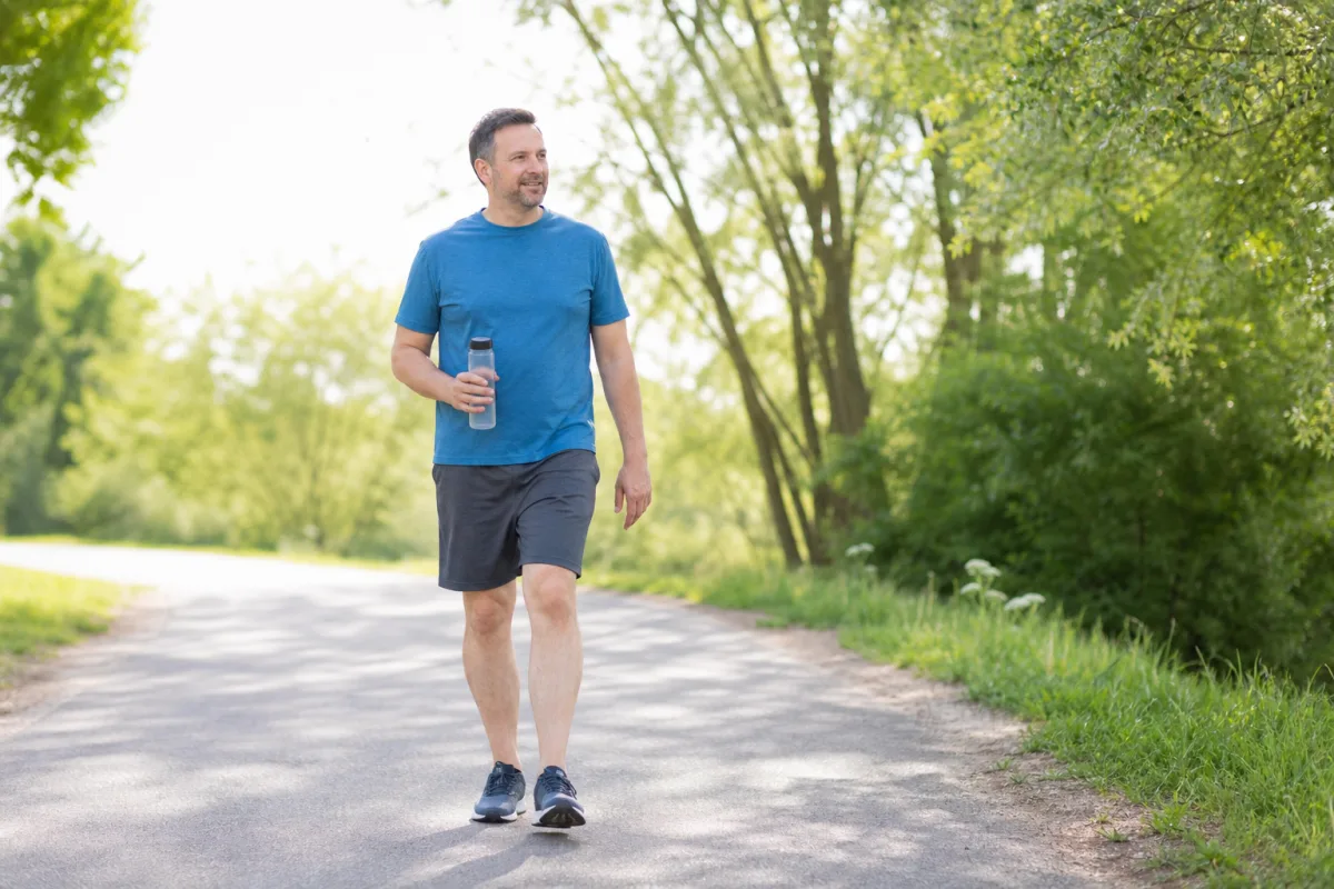 A man walking on a bright outdoor path carrying a water bottle, trees and morning light in the background