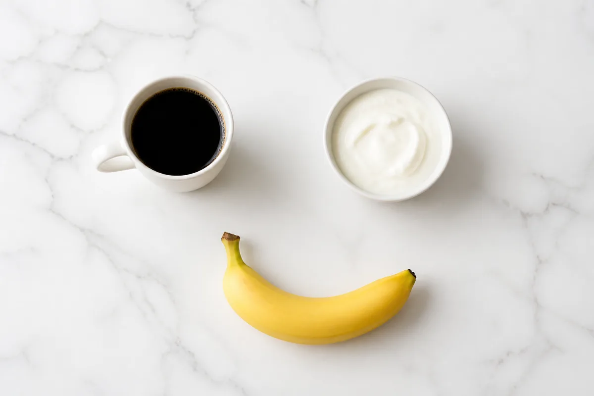 A white mug of black coffee, a bowl of plain Greek yogurt, and a banana on a bright marble surface