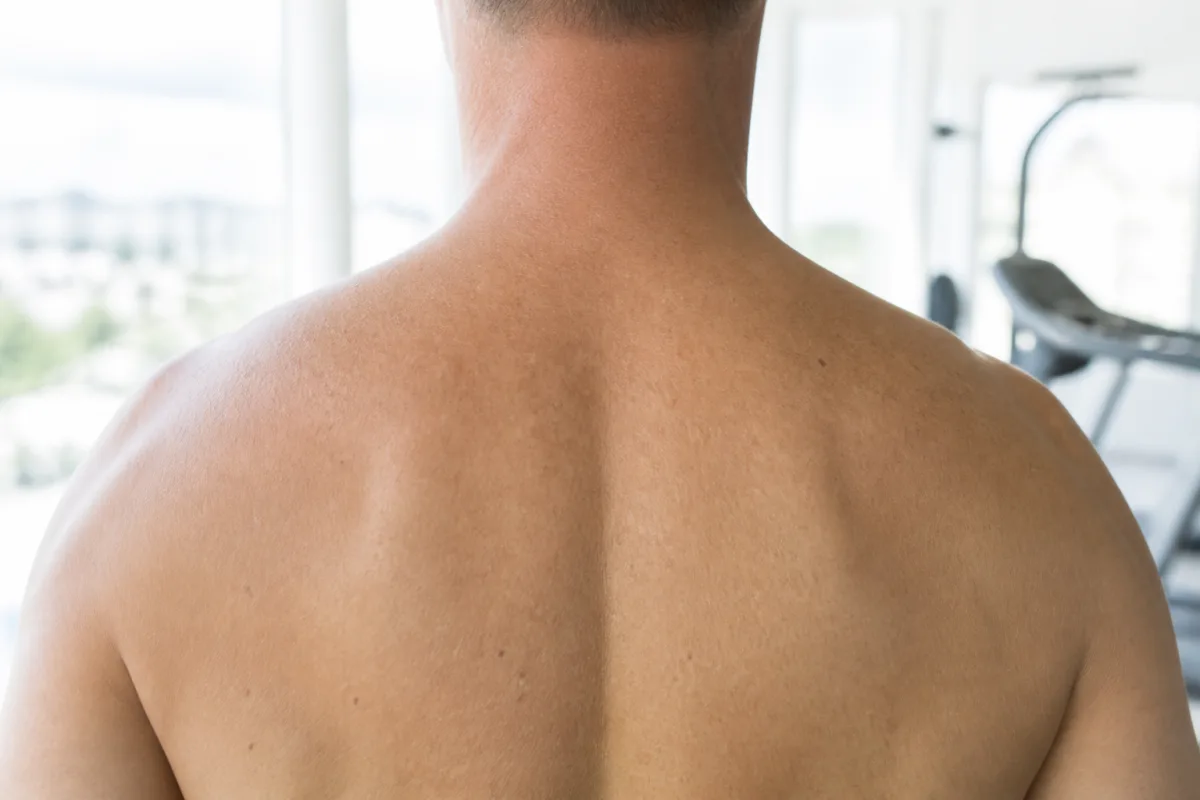 Back view of a man's shoulders and upper back at a bright gym window, showing the upper trapezius region between neck and shoulder