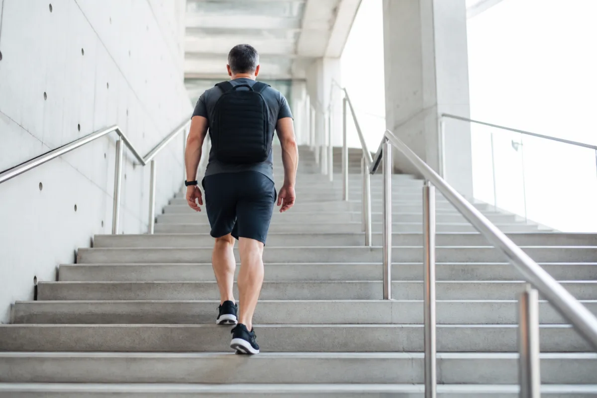 A man in his forties climbing a bright outdoor staircase wearing a weighted backpack